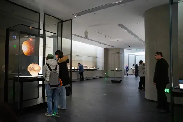 Interior de uma galeria moderna do Museu de Xangai, onde visitantes observam artefatos históricos em vitrines de vidro iluminadas. Em primeiro plano, duas mulheres de costas analisam um grande vaso de cerâmica antiga exposto individualmente, enquanto ao fundo outros visitantes exploram uma longa vitrine embutida na parede cinza minimalista. A iluminação é focada nas obras, criando um ambiente contemplativo e sofisticado.
