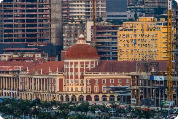 Vista urbana de Luanda, Angola, focando no edifício histórico do Banco Nacional de Angola com sua cúpula vermelha e fachada rosa e branca. Ao redor, misturam-se prédios modernos em construção e guindastes, sob uma luz quente de fim de tarde.
