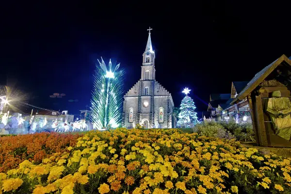 Igreja Matriz São Pedro, em Gramado, iluminada para o Natal, com árvores decoradas e jardim florido em primeiro plano.