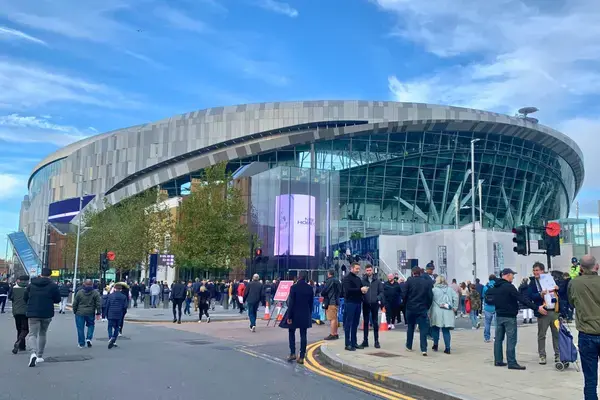 Vista externa do Tottenham Hotspur Stadium em Londres, com fachada moderna em metal e vidro e torcedores circulando na entrada principal em dia de evento.