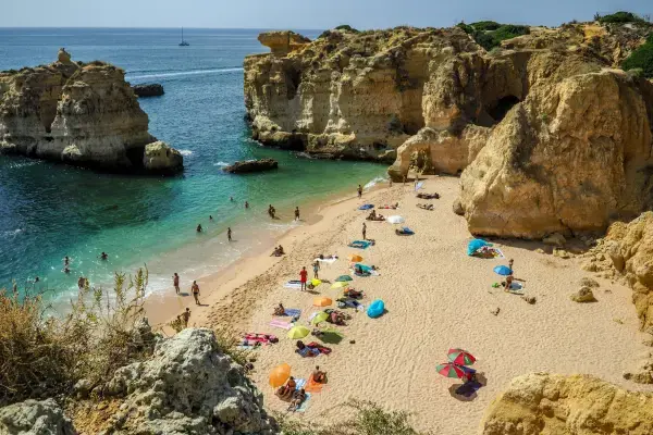 Praia de areia clara e falésias douradas com turistas e guarda-sóis coloridos no Algarve, Portugal.