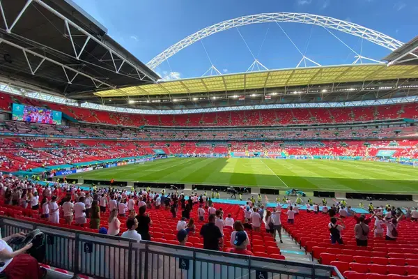 Vista interna do Wembley Stadium, na Inglaterra, com arquibancadas vermelhas, torcedores nas cadeiras e no setor inferior, gramado verde impecável ao centro e cobertura metálica arqueada sobre a arena em dia de jogo.