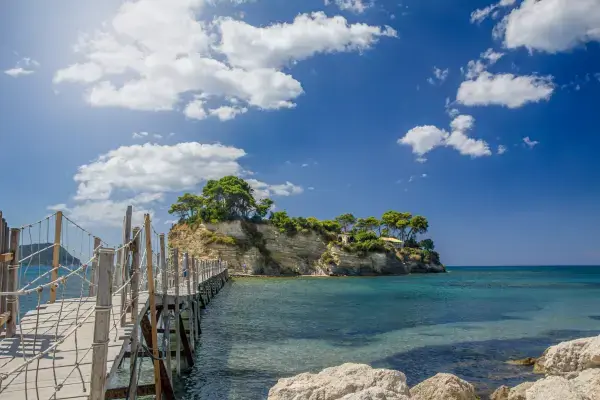 Passarela de madeira sobre águas cristalinas em Zakynthos, com ilhota rochosa coberta por vegetação ao fundo e céu azul.