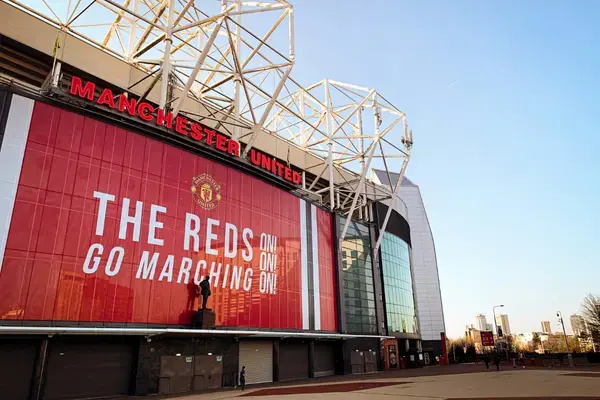 Fachada do Old Trafford, estádio do Manchester United, com painel vermelho escrito “The Reds Go Marching On” e estrutura metálica branca no topo.