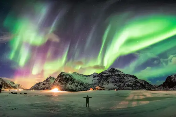 Vista panorâmica de uma Aurora Boreal intensa com faixas de luz verde, roxa e amarela cruzando todo o céu noturno. Abaixo, montanhas cobertas de neve e pequenas casas iluminadas cercam uma planície gelada onde se vê a silhueta de uma pessoa com os braços abertos.
