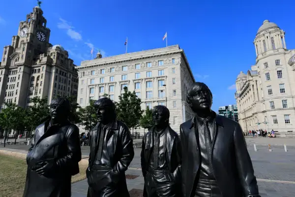 Estátuas em bronze dos quatro integrantes dos Beatles em Liverpool, posicionadas em uma praça com prédios históricos ao fundo.