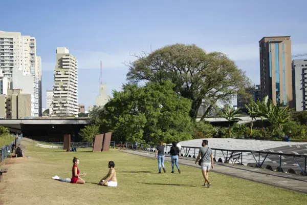 Espaço gramado com pessoas sentadas e caminhando no Centro Cultural São Paulo, cercado por prédios altos ao fundo. A estrutura do centro cultural aparece parcialmente à direita, com árvores grandes ao redor.