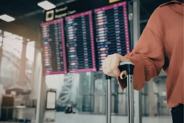 Mão segurando mala, ao fundo, painel de voos em aeroporto com horários de embarque