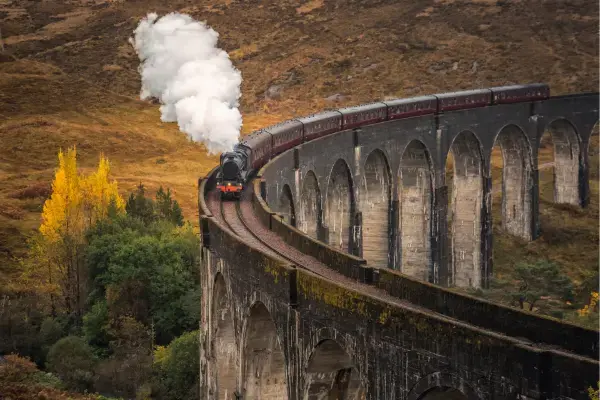 Trem a vapor atravessando o viaduto curvo de Glenfinnan, rodeado por colinas e vegetação outonal na Escócia.