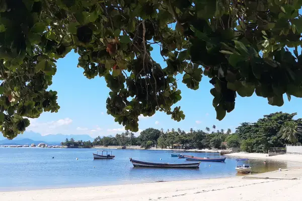 Praia calma com areia clara, barcos ancorados no mar e árvores ao fundo. A imagem foi tirada na Ilha de Paquetá, no Rio de Janeiro.