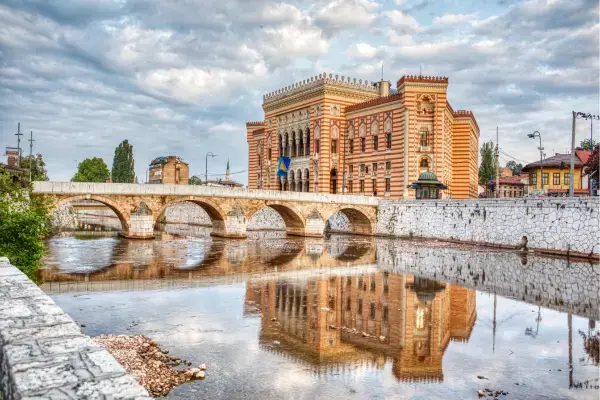 Prédio histórico de fachada ornamentada em tons de vermelho e bege, refletido nas águas de um rio. À frente, há uma ponte de pedra com arcos que atravessa o rio. O céu está parcialmente nublado.