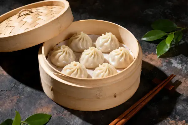 Cesta de bambu aberta contendo sete bolinhos Xiao Long Bao cozidos no vapor. A tampa da cesta está ao lado, junto com hashis de madeira sobre um suporte. A cena é decorada com folhas verdes em um fundo escuro e rústico, com iluminação quente.
