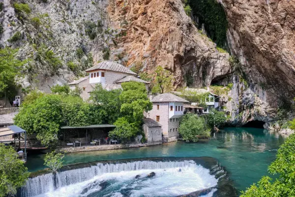Casas brancas ficam ao lado de um penhasco, perto de um rio de água azul que sai de dentro da rocha, formando pequenas quedas d’água.