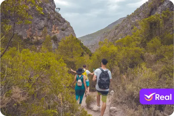 Esta é uma fotografia vibrante e imersiva que captura um momento de aventura e exploração na natureza. A imagem retrata três jovens adultos caminhando por uma trilha estreita em um vale ou cânion, cercados por uma vegetação densa e paisagens rochosas. A perspectiva da foto, tirada por trás do grupo, convida o espectador a se sentir parte da jornada. Os Personagens Principais O foco da imagem está em três pessoas, vistas de costas, que parecem ser um grupo de amigos desfrutando de uma caminhada: À direita e em primeiro plano, um jovem veste uma bermuda verde-clara e carrega uma mochila preta com detalhes em branco. Em sua mão esquerda, ele segura o que parece ser uma garrafa de água ou um celular. No centro, parcialmente encoberto, um segundo jovem usa uma camiseta branca. Seus braços estão abertos para os lados, em um gesto que transmite liberdade e uma conexão com o ambiente ao redor. À esquerda, uma jovem veste um macacão ou vestido longo em um tom de verde-azulado. Ela usa um chapéu de palha de abas largas, ideal para proteção solar, e também carrega uma pequena mochila clara. Seu braço esquerdo está estendido, talvez para se equilibrar ou simplesmente para tocar a vegetação. A disposição do grupo em fila indiana sugere que a trilha é estreita e selvagem, reforçando o sentimento de aventura. O Ambiente e a Paisagem A cena se passa em um ambiente natural e robusto: A Trilha: Um caminho de terra batida e poeirenta serpenteia pelo cenário, sendo o único indício de passagem humana na vegetação densa. A Vegetação: A flora é abundante e dominante na imagem, com arbustos e árvores de médio porte preenchendo todo o vale. A folhagem tem um tom predominante de verde-oliva e amarelado, sugerindo um clima quente e talvez mediterrâneo. O Relevo: Em ambos os lados da trilha, elevam-se encostas íngremes e rochosas, características de um cânion. As formações rochosas cinzentas contrastam com o verde da vegetação. O Céu: O céu está encoberto por nuvens, o que cria uma iluminação suave e difusa, sem sombras fortes, conferindo um aspecto natural e realista à cena. Composição e Atmosfera A fotografia transmite uma forte sensação de aventura, amizade e descoberta. A paleta de cores, dominada por tons terrosos e verdes, é natural e convidativa. A composição, que segue o grupo em sua caminhada para o interior da paisagem, cria uma narrativa de exploração e convida o espectador a imaginar o que eles encontrarão pela frente. Elemento Gráfico No canto inferior direito da imagem, há um elemento gráfico sobreposto: um retângulo roxo com cantos arredondados. Dentro dele, é possível ver um ícone de "check" (✓) branco e parte do nome "Real", identificando a imagem como material promocional da Real Seguro Viagem.
